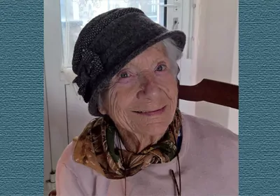 Older woman with hat and pink blouse smiling at camera.